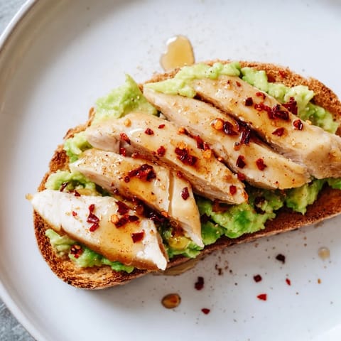 Creamy smashed avocado and grilled chicken slices are piled high on golden, crusty sourdough toast for a hearty, savory-sweet bite.  