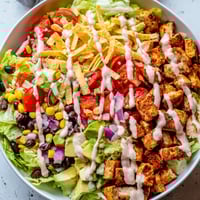 A vibrant Chicken Taco Chopped Salad in a white bowl, loaded with seasoned chicken, crisp romaine, black beans, corn, diced avocado, and crushed tortilla chips, drizzled with creamy salsa ranch dressing.  